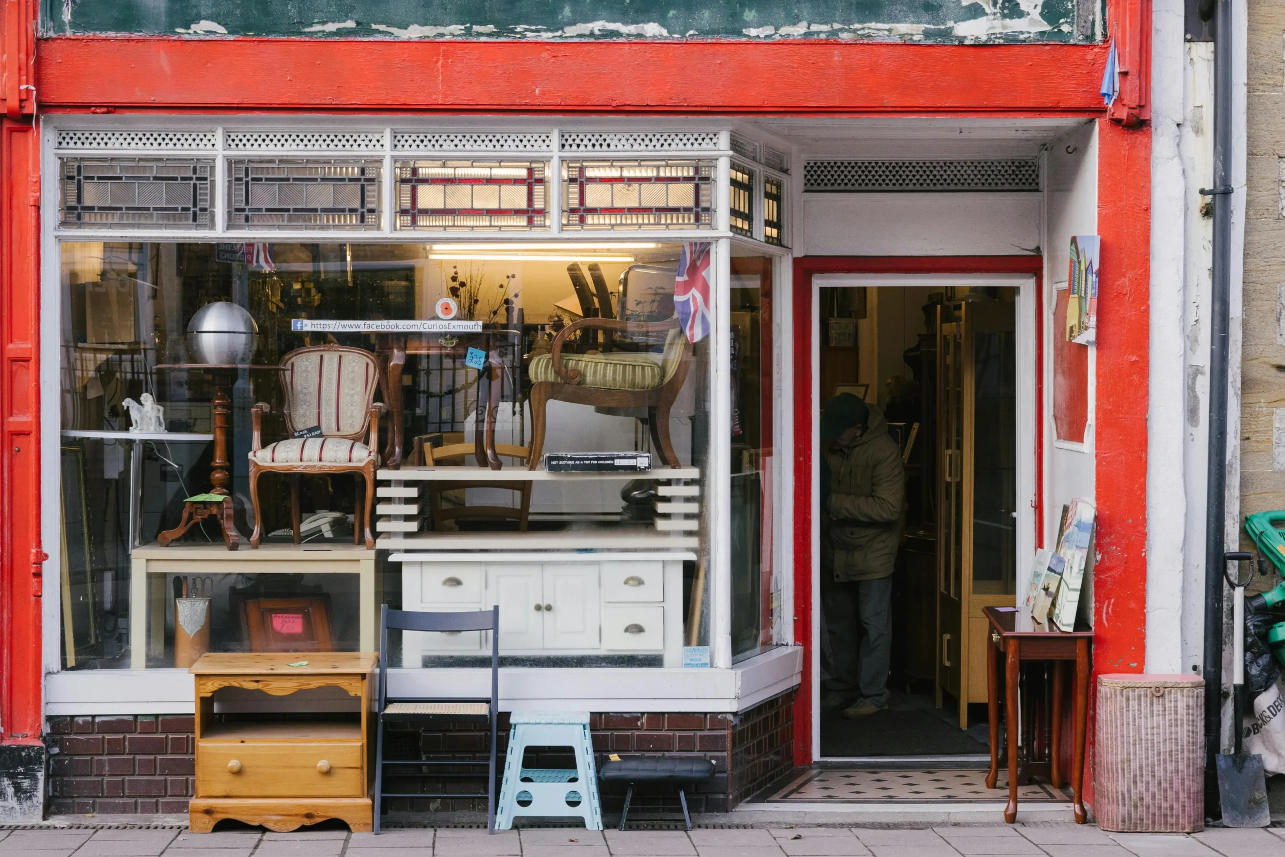 Front view of an antique shop with a red frame, showing vintage furniture like chairs and tables in the window, and a person inside browsing items.