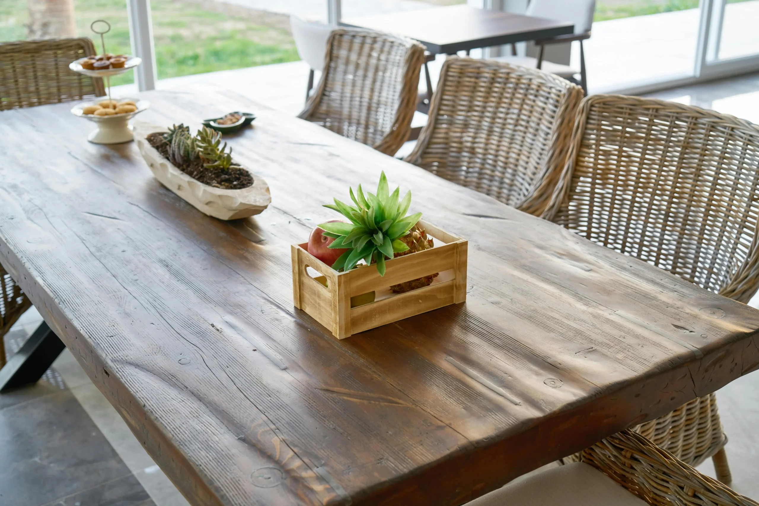 Rustic-style wooden table with wicker chairs, decorated with a succulent centerpiece, fruit tray, and a plate of snacks.