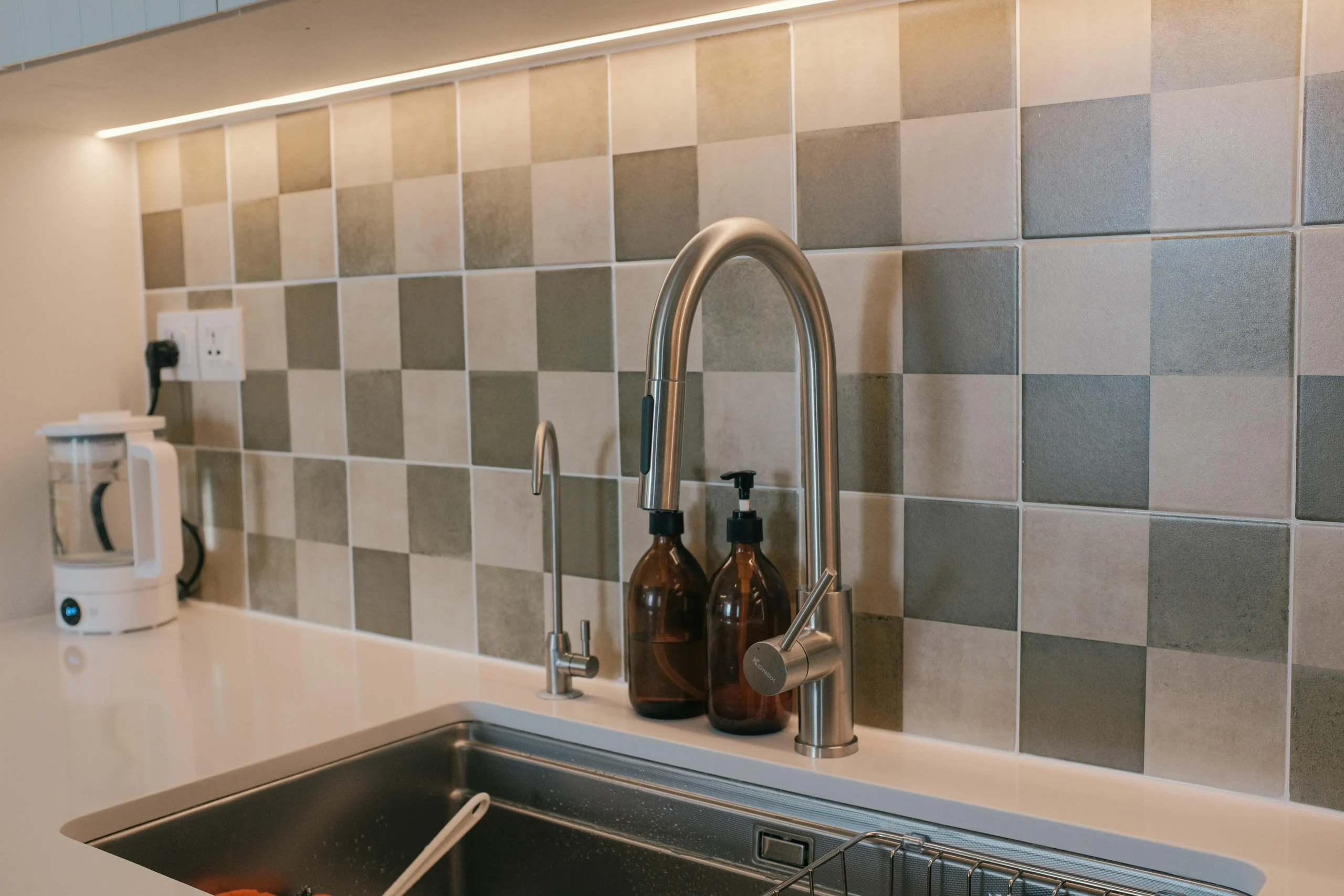 Fregadero de cocina de acero inoxidable con un grifo metálico alto, dos dispensadores de jabón marrones, una pared de azulejos en tonos grises y beige, y una cafetera blanca sobre la encimera.
