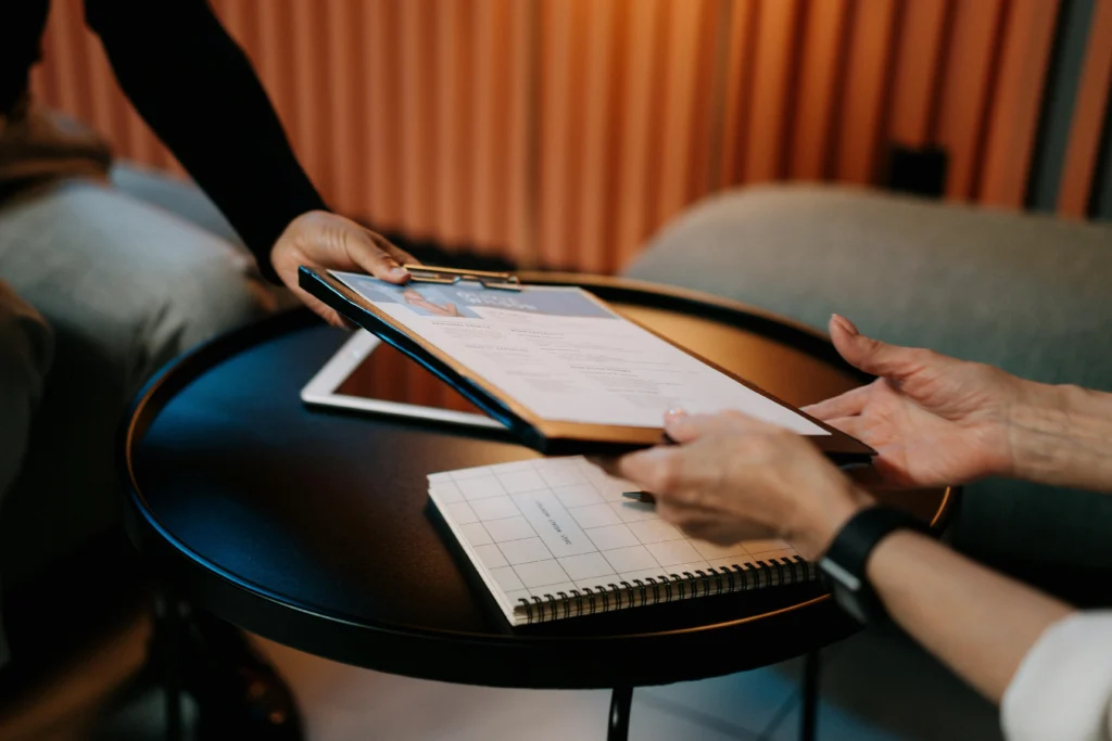 A person handing a document to another across a table, next to a notebook and a tablet.