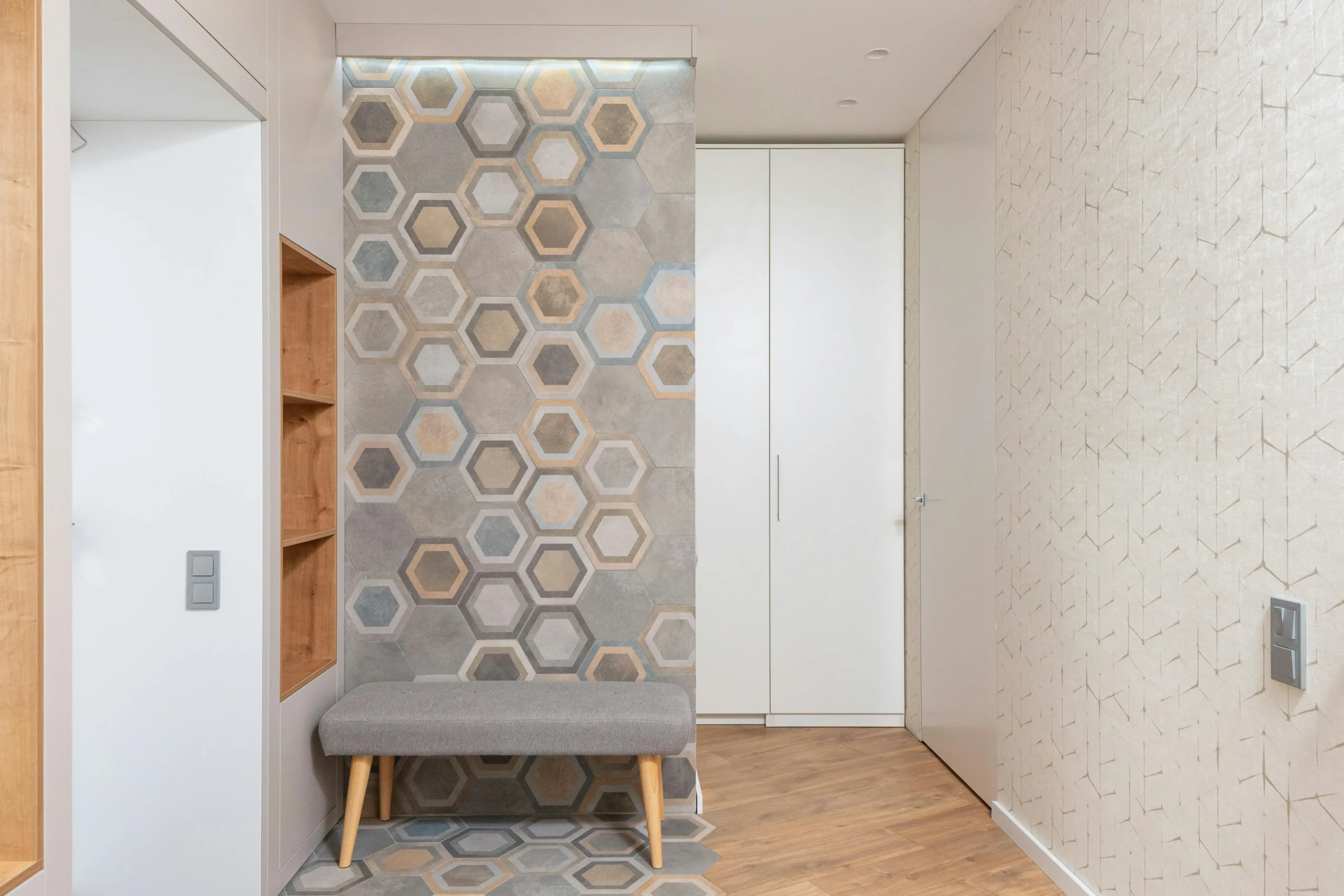 Vista d’un rebedor modern amb paret decorativa de rajoles hexagonals en tons neutres, banc entapissat en gris clar amb potes de fusta, armari blanc encastat i terra de parquet.