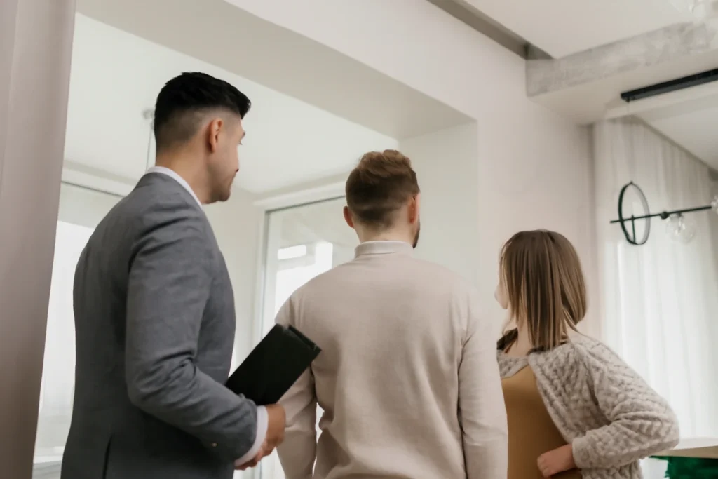 Three people—a man in a suit and two clients—are viewing an apartment in Barcelona. They are standing in a bright, modern room, with the man holding a clipboard and the clients looking at each other as they interact.