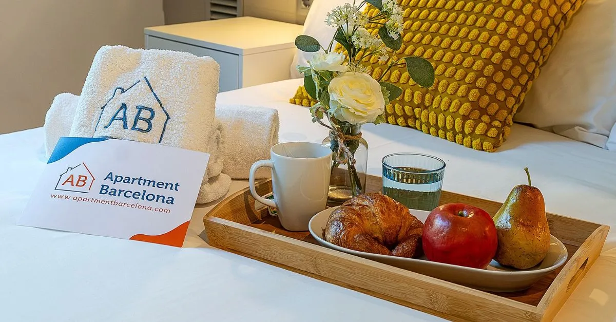 Tray with croissant, fruit, coffee and water next to fresh flowers and towels with the AB Apartment Barcelona logo as a welcome kit for guests.