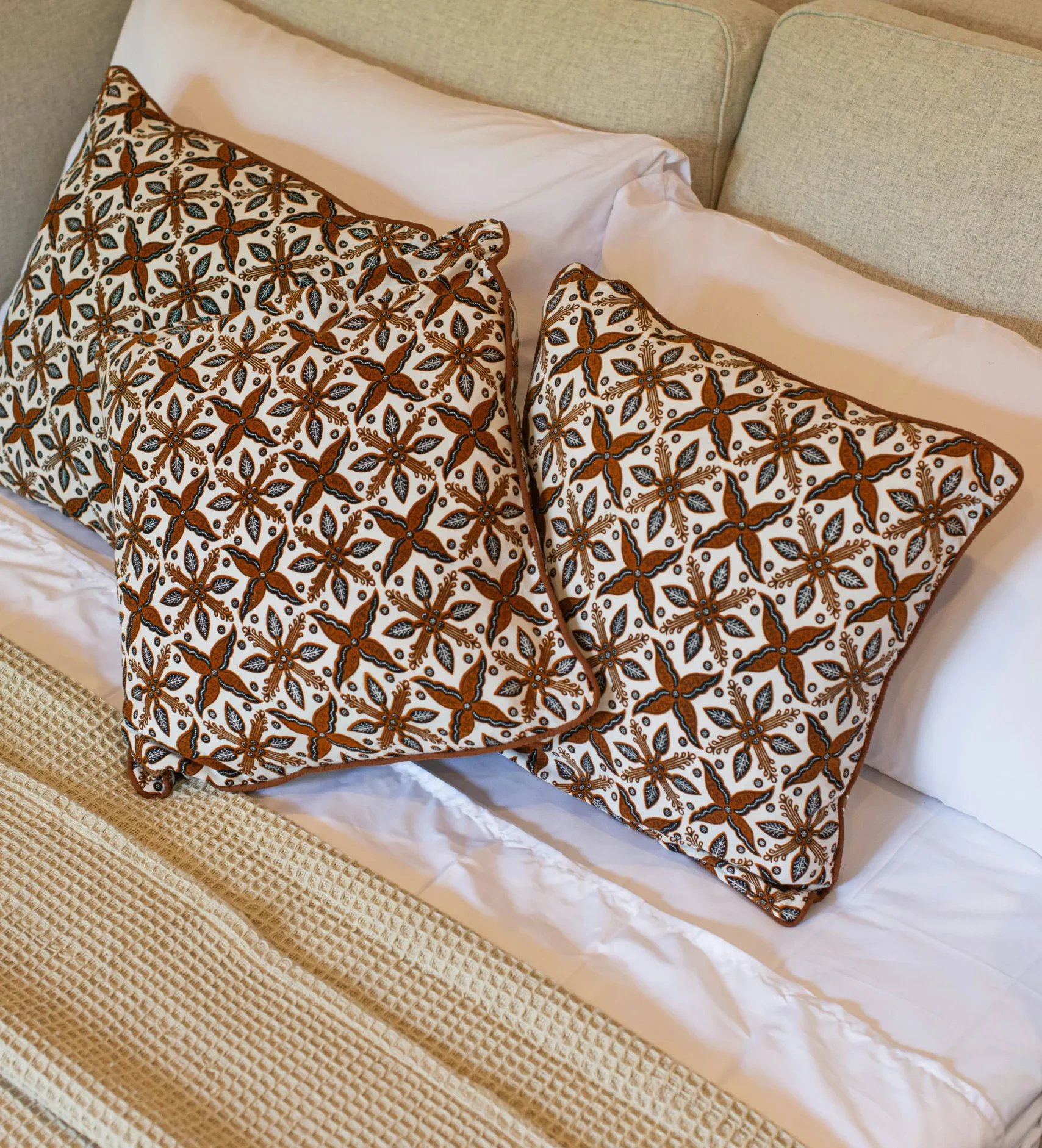 Cama con cojines decorativos de estampado marrón y blanco con diseño floral geométrico, acompañada de manta beige de punto.