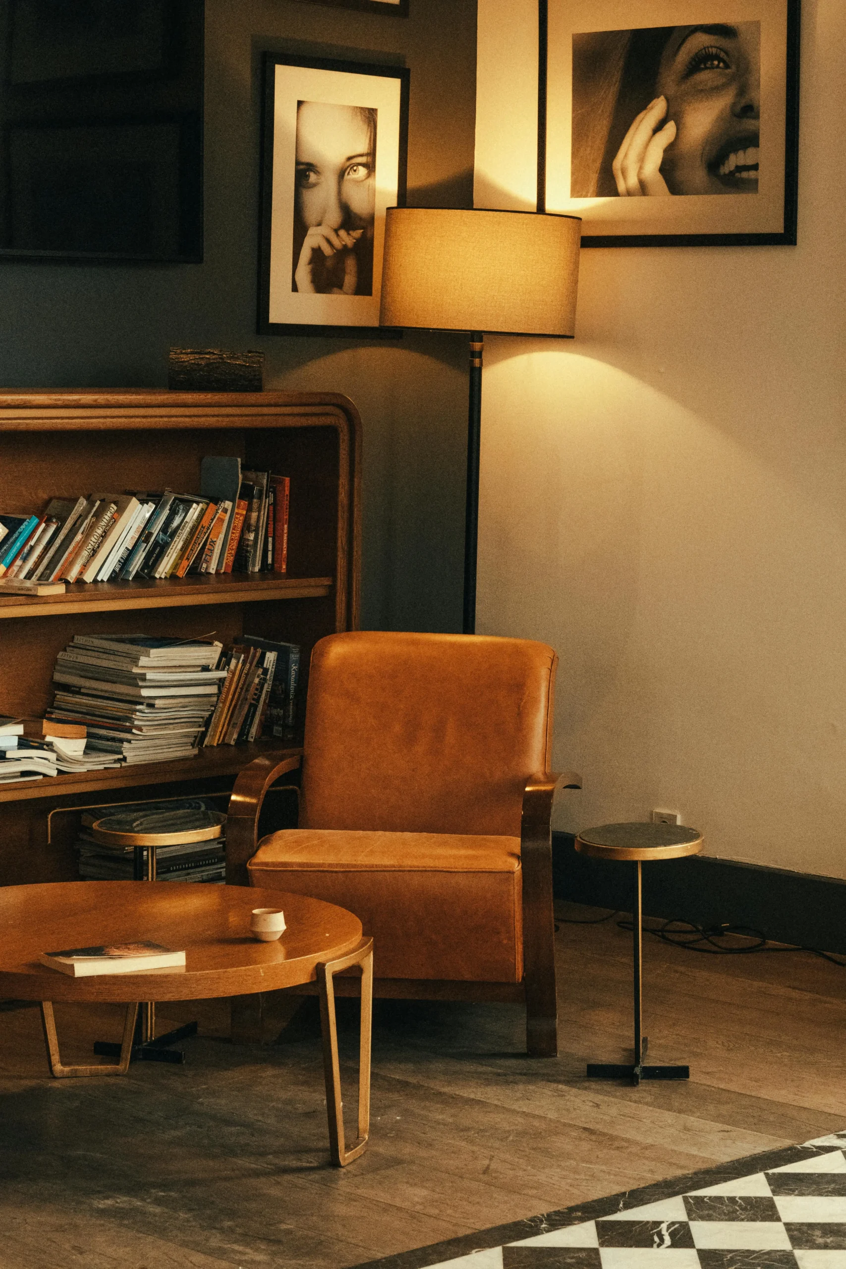 Sillón de cuero marrón junto a una lámpara de pie encendida, con estantería de libros y mesa de madera en un salón acogedor de estilo vintage 