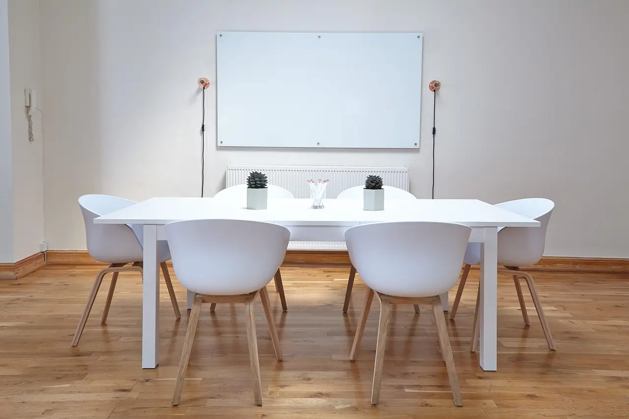 View of a meeting room with a white rectangular table, six modern white chairs with wooden legs, two small decorative plants, and a whiteboard on the wall.