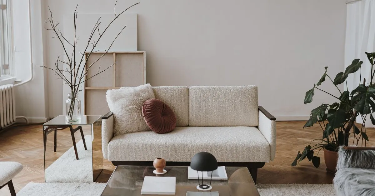 Salón minimalista con sofá beige, mesa de vidrio, cojines decorativos y plantas que aportan un ambiente natural y acogedor
