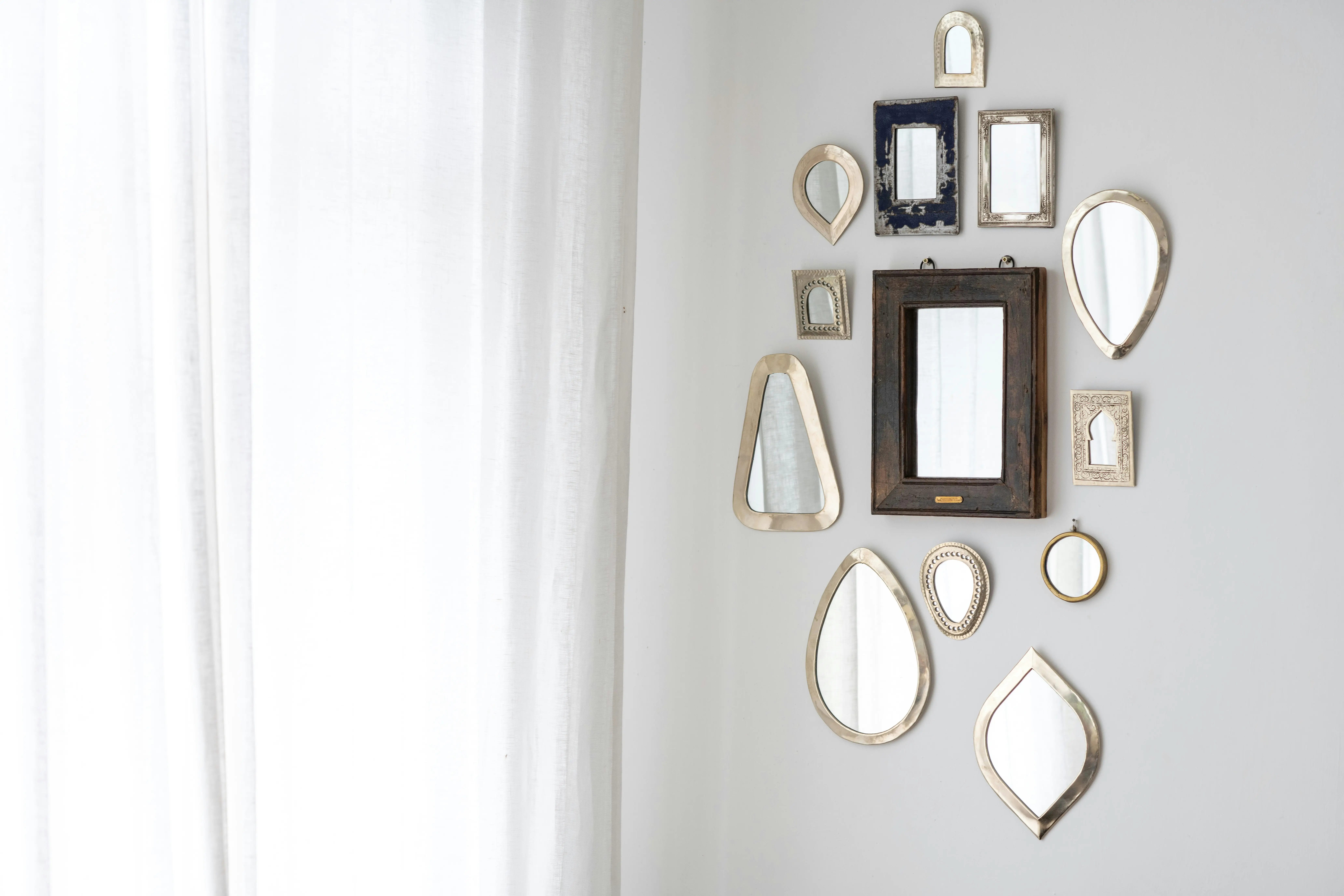 Group of decorative mirrors with metal and wooden frames on a white wall next to a light curtain.