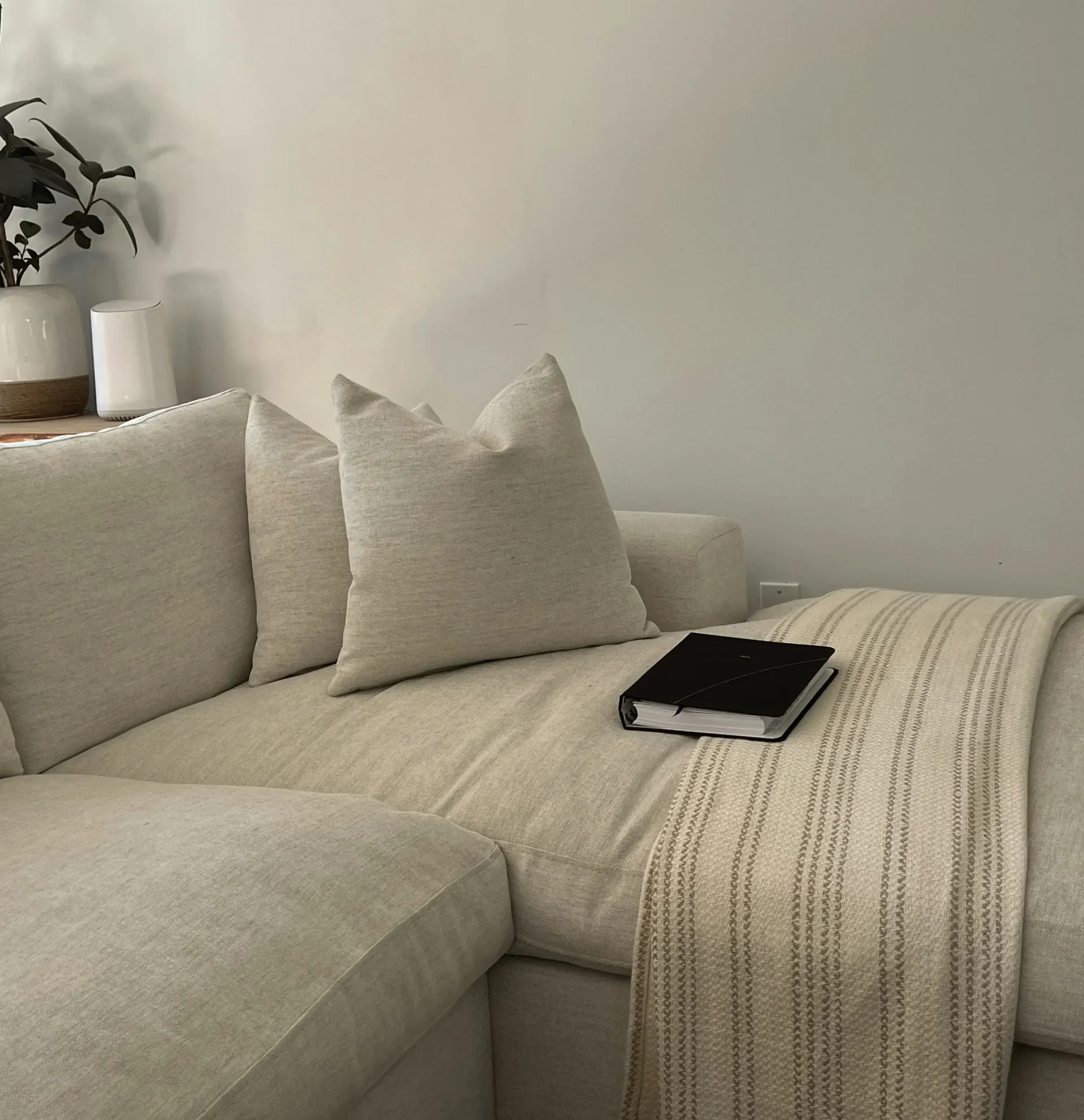 Beige-toned sofa with cushions, a knitted throw, and a black book on the seat in a minimalist setting.