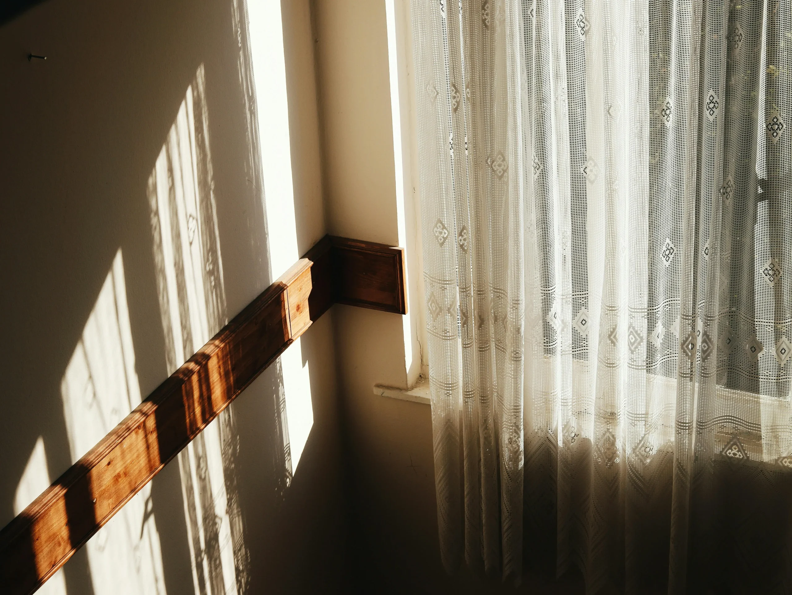 Detail of a window with a white sheer curtain letting natural light through, casting soft shadows on the wall for a warm and cozy touch.