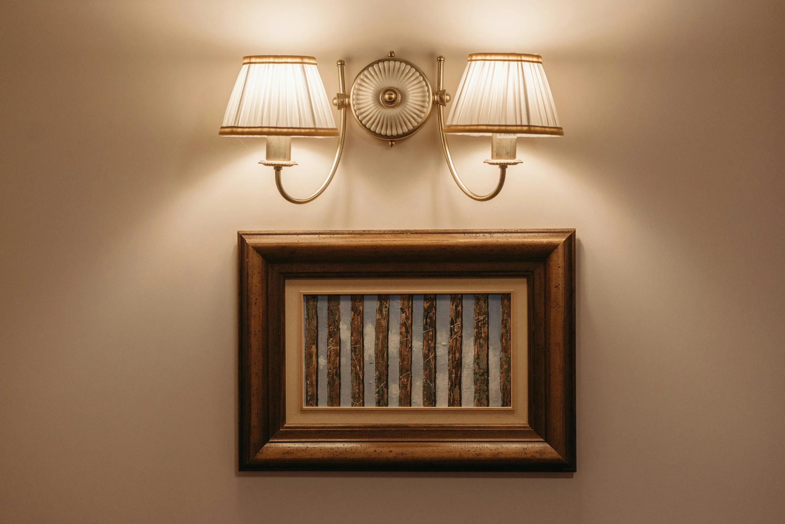 Interior wall decorated with a framed classic-style painting, illuminated by a wall sconce with two beige-shaded lamps that create a warm and welcoming atmosphere.