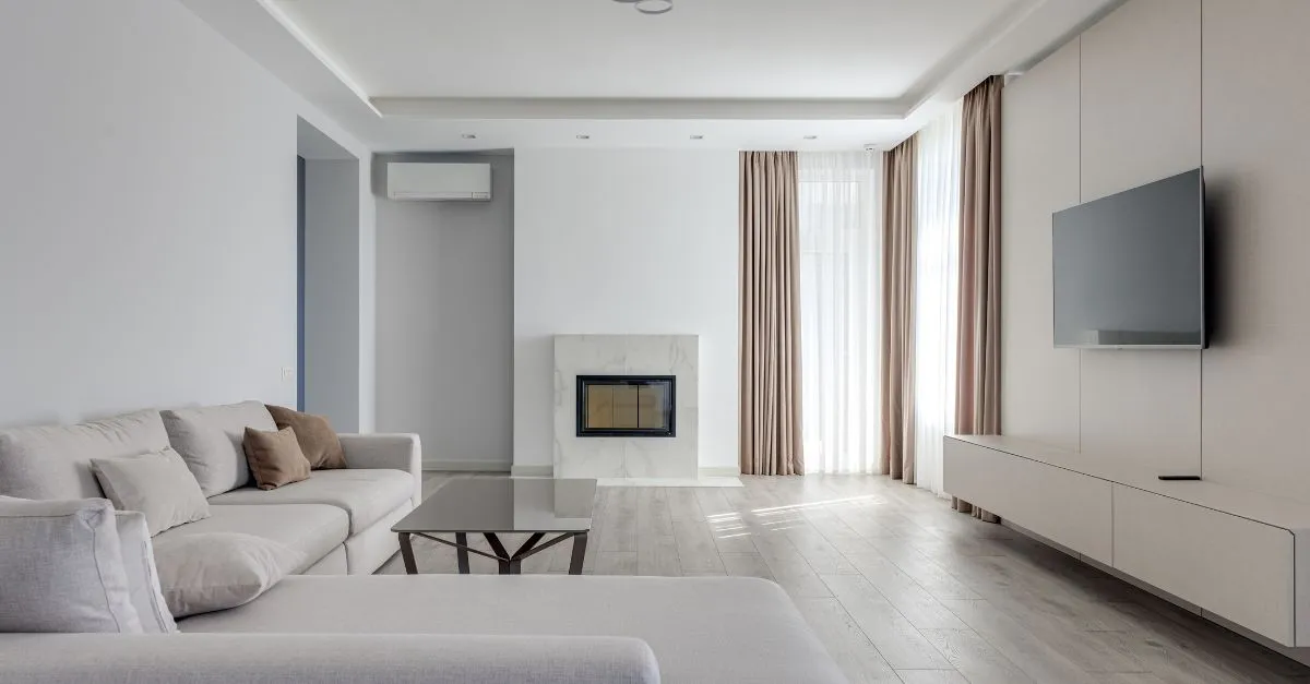 Sala de estar decorada en tonos neutros con sofá claro, chimenea moderna, cortinas beige y televisión de pared