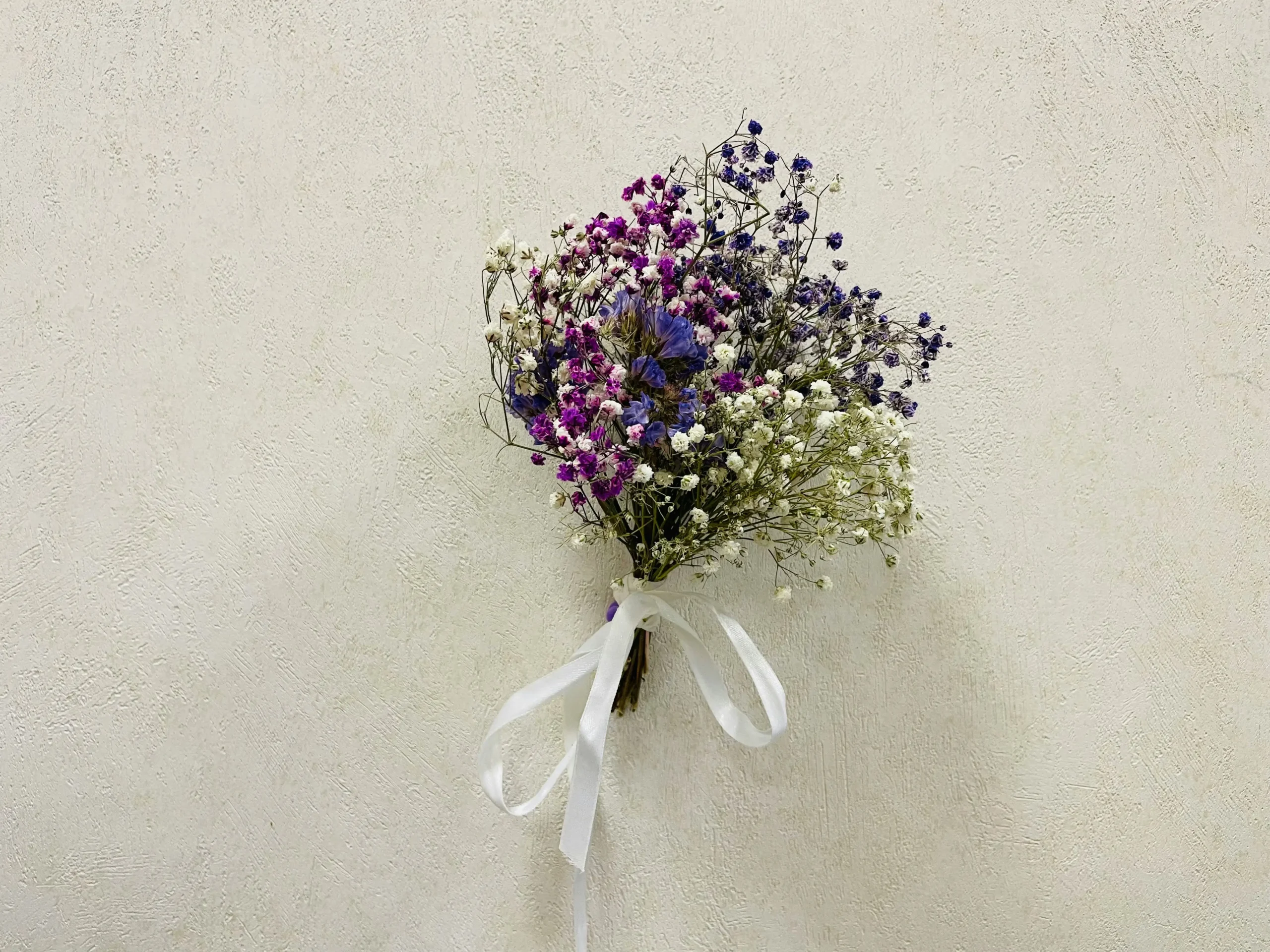 Ramillete de flores secas en tonos morados, blancos y azules, atado con un lazo blanco y colgado sobre una pared de color claro con textura. 