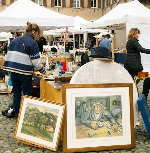 Parada d’un mercat a l’aire lliure amb quadres emmarcats recolzats a terra, taules amb objectes decoratius i diverses persones explorant peces d’art i antiguitats.