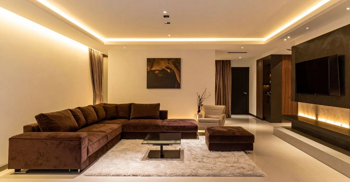 Spacious contemporary living room decorated in brown tones with an L-shaped sofa, beige rug, and warm indirect lighting that creates a cozy, elegant atmosphere.