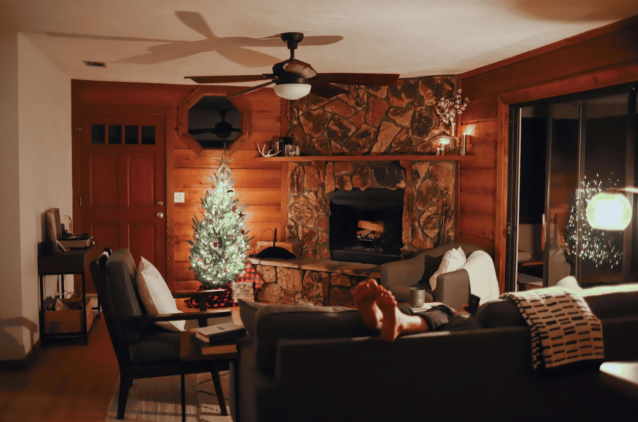 Image of a rustic-style living room with wooden walls, a small lit Christmas tree, a stone fireplace, cozy furniture, and a person relaxing on the sofa with a cup nearby.