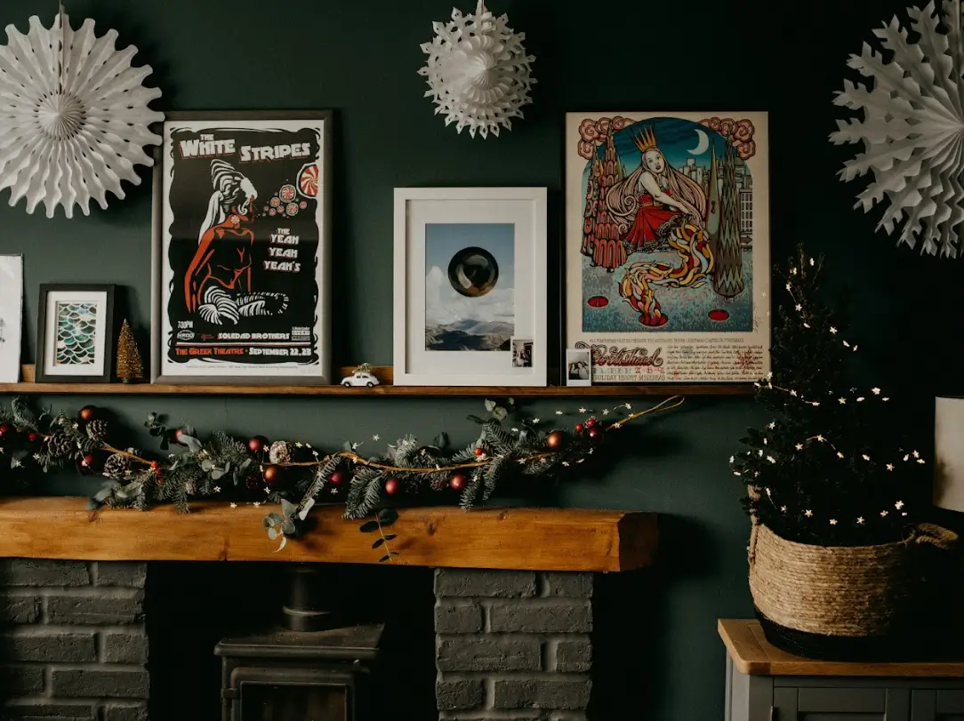 Image of a fireplace decorated with a Christmas garland made of branches, pinecones, and red ornaments. On the mantel, there are framed posters and prints, along with a small lit Christmas tree placed in a basket.