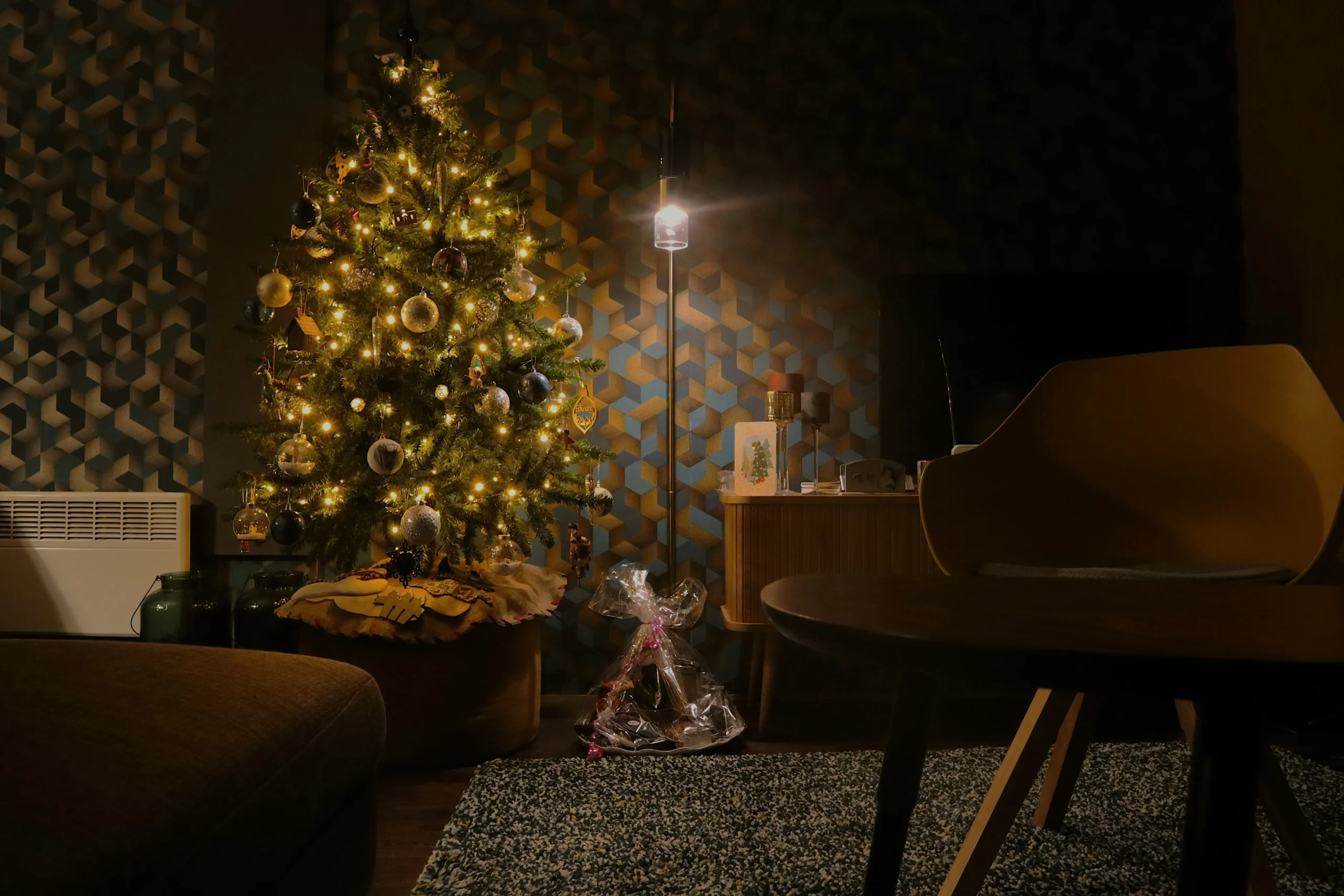 Image of a living room with a Christmas tree decorated with lights and gold ornaments, a lit lamp, a sideboard with decorative items, and a gift wrapped in cellophane placed on a rug.