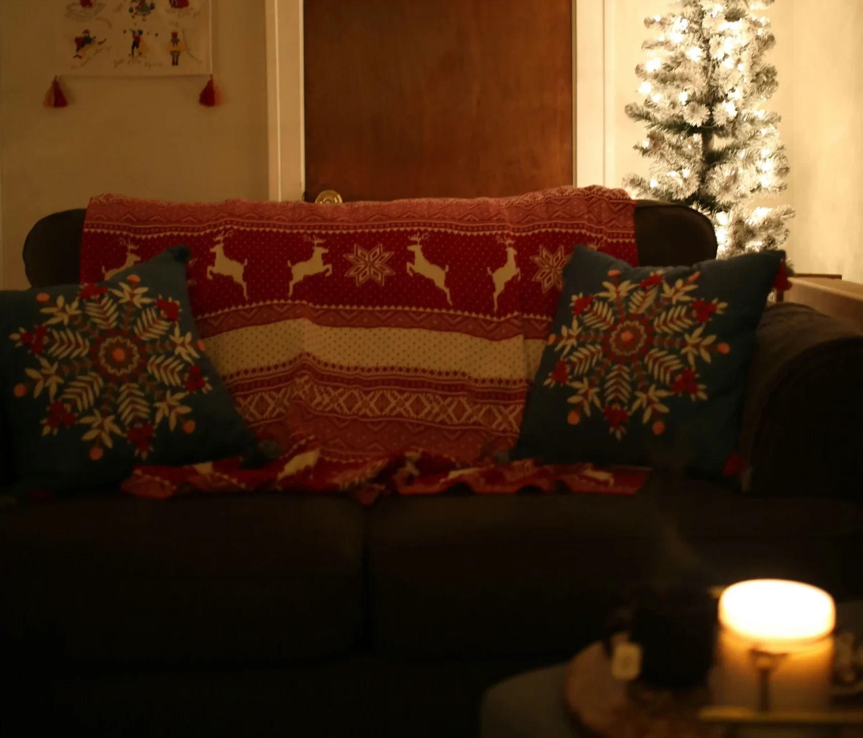 Image of a living room decorated for Christmas with a sofa covered by a red winter-themed throw, blue cushions with festive designs, a small lit Christmas tree, and a candle on a table.