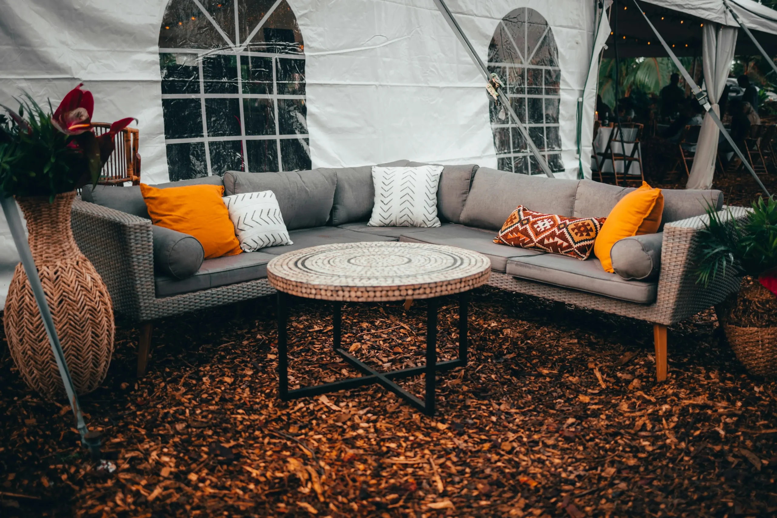 Image of an outdoor space with a wicker corner sofa and cushions in white, orange, and ethnic tones. In the center, there is a round decorative table on a wood-chip floor, surrounded by plants and boho-style elements.