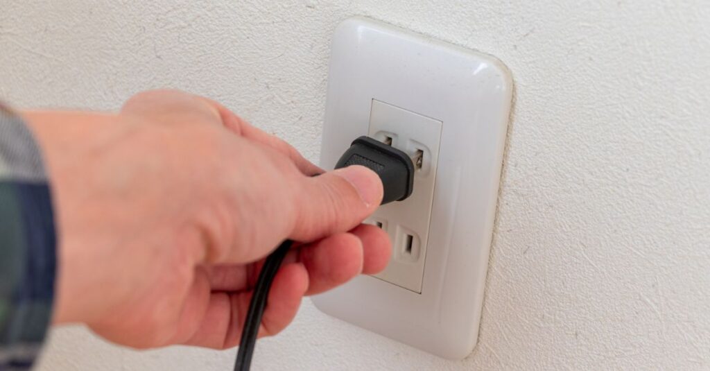 Primer pla d’una mà connectant un cable a un endoll de paret blanc. L’enfocament se centra en l’acció d’introduir el connector a la presa de corrent.