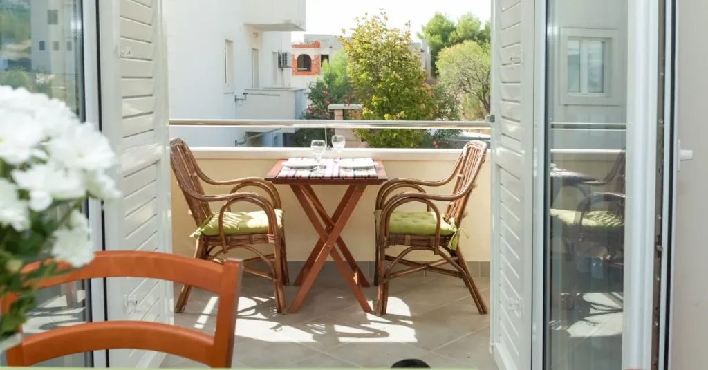 Vista d'una terrassa amb una taula de fusta i dues cadires de vímet, amb coixins verds. La taula està parada amb plats i copes, i al fons s’hi pot veure una planta i edificis propers, gaudint d’un ambient lluminós i relaxant.