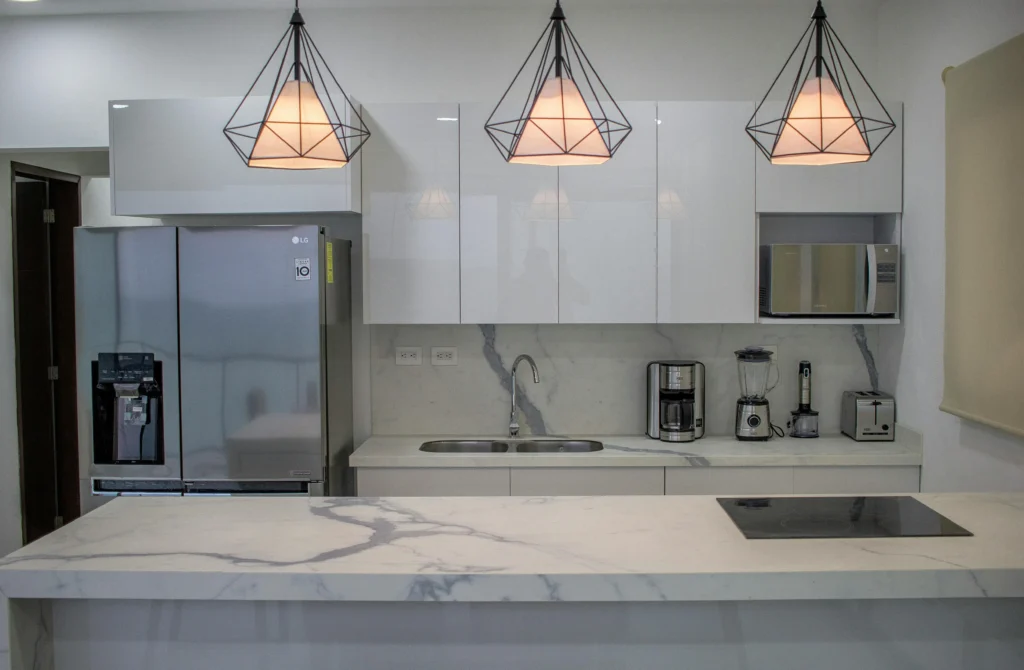Cocina moderna de diseño minimalista con isla de mármol blanco, armarios lacados en blanco, frigorífico de acero inoxidable, placa de inducción y lámparas colgantes geométricas.
