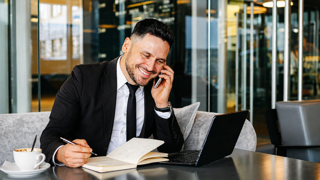 Profesional sonriente realizando una llamada de negocios mientras toma notas en una libreta y trabaja con un portátil en un entorno moderno y luminoso.