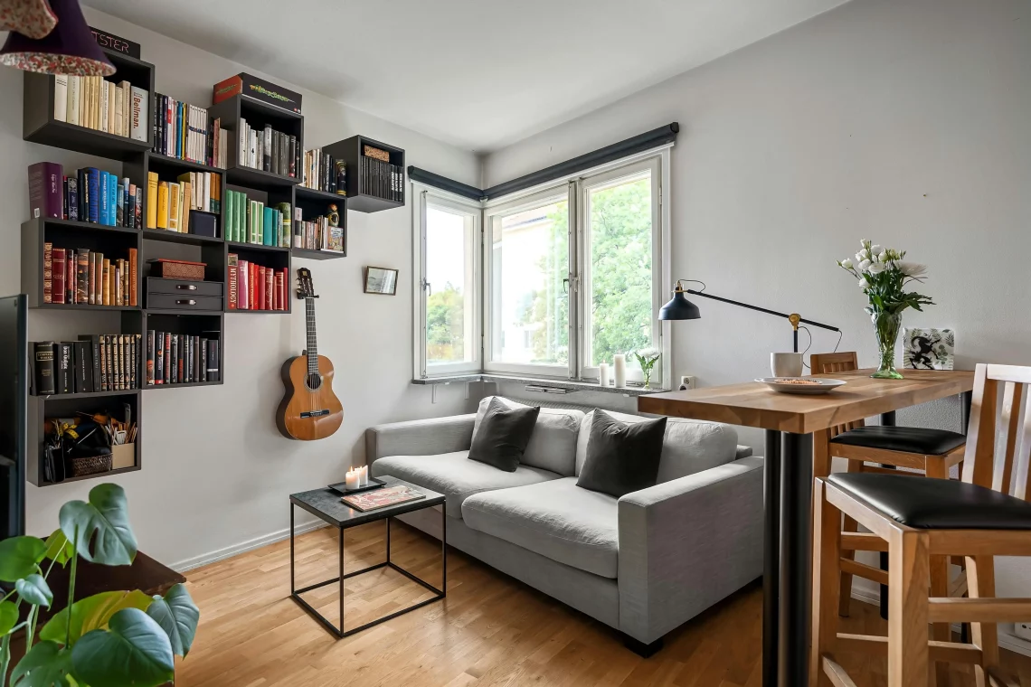 Sala de estar pequeña y bien decorada con una estantería llena de libros, una guitarra en la pared, un sofá gris y una mesa alta de madera, mostrando cómo optimizar la decoración en un piso pequeño con un presupuesto reducido para aumentar su valor