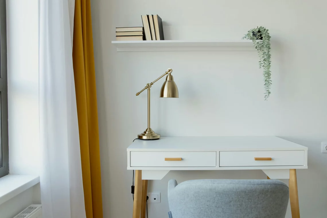 Vista de un escritorio blanco de estilo moderno, con una lámpara dorada y una silla gris a su lado.