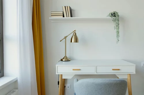 Vista de un escritorio blanco de estilo moderno, con una lámpara dorada y una silla gris a su lado.