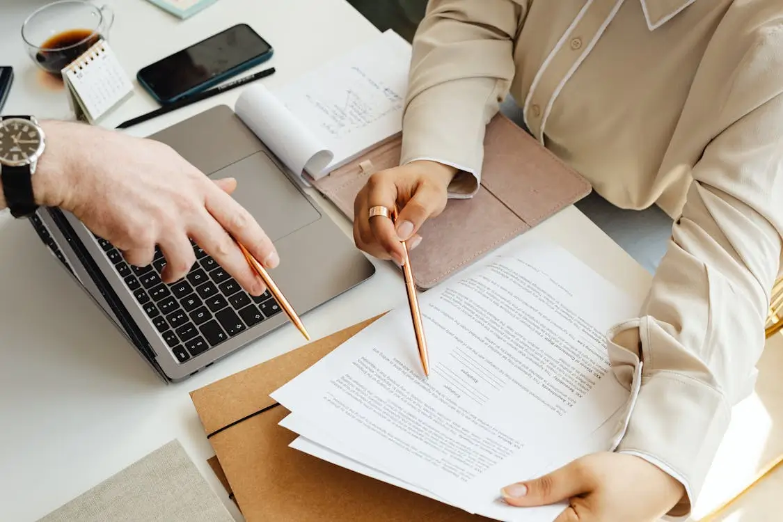 Dos personas revisando documentos y contratos sobre la mesa, con un ordenador portátil abierto