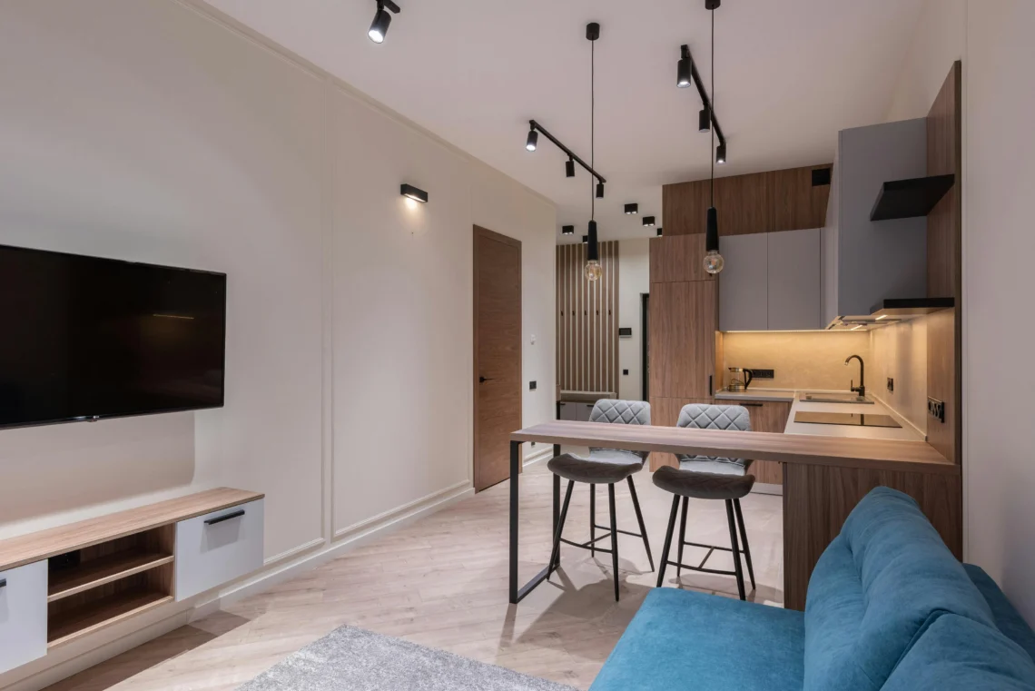 Apartment interior with contemporary design, wooden kitchen with an island and cushioned stools, blue sofa, flat-screen TV, and ceiling spotlights for focused lighting