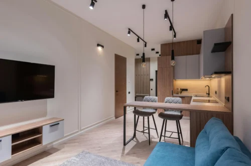 Apartment interior with contemporary design, wooden kitchen with an island and cushioned stools, blue sofa, flat-screen TV, and ceiling spotlights for focused lighting