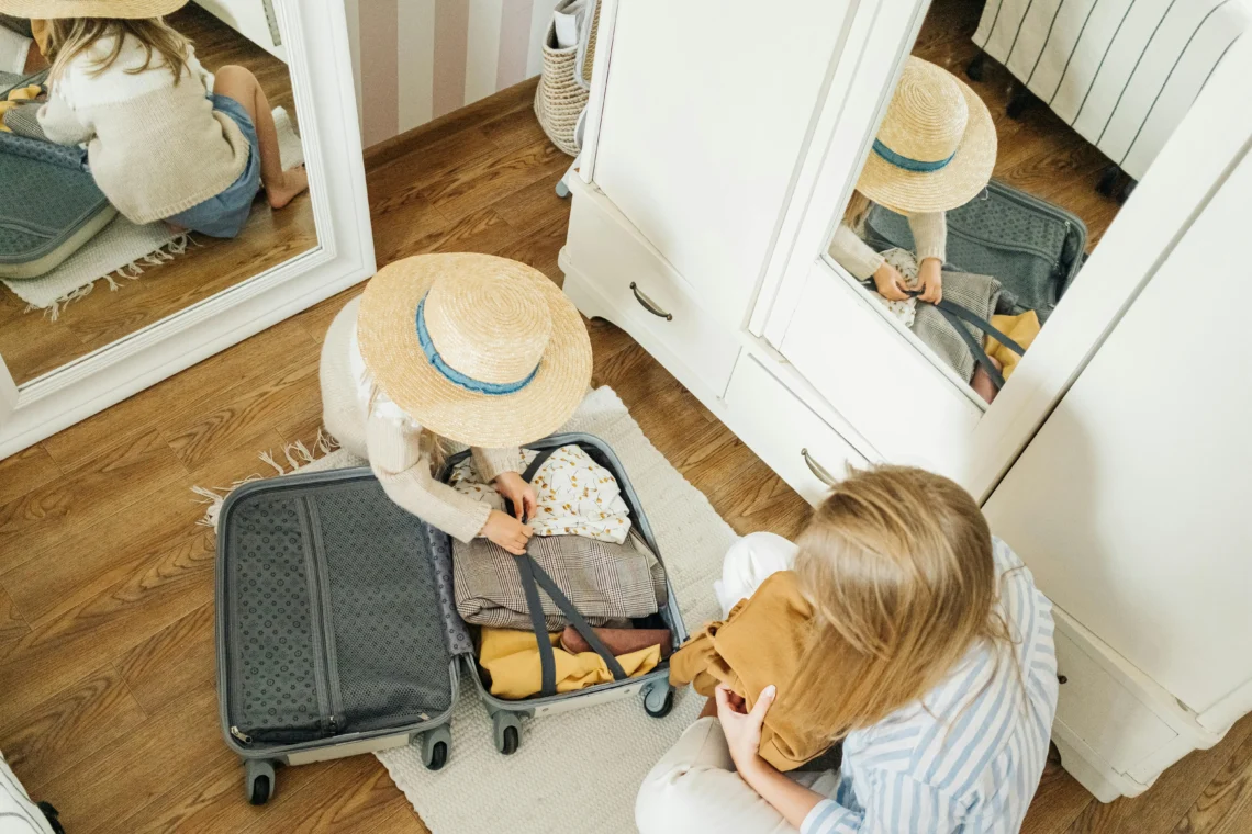 Top-down view of a girl wearing a straw hat closing a suitcase next to her mother, in a room with wooden flooring, a light-colored rug, and white mirrored wardrobes.