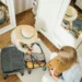 Top-down view of a girl wearing a straw hat closing a suitcase next to her mother, in a room with wooden flooring, a light-colored rug, and white mirrored wardrobes.