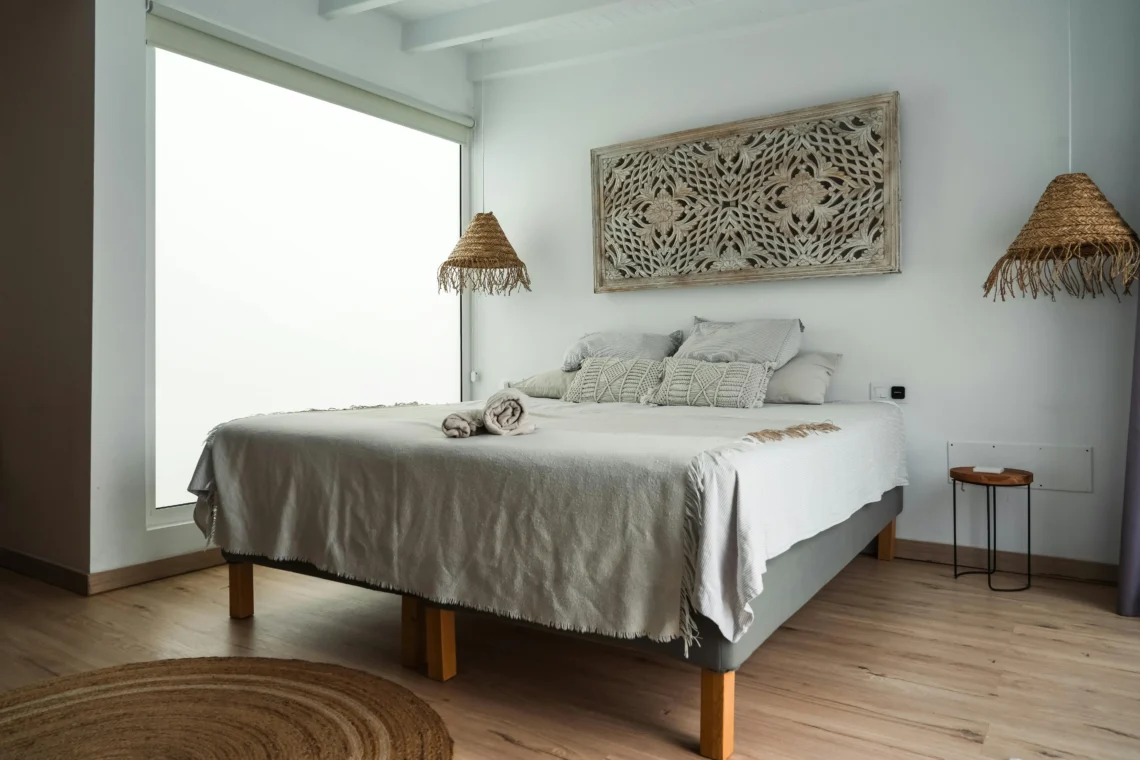 Minimalist bedroom with neutral tones and natural textures, featuring hanging wicker lamps, macramé cushions, and a wooden side table.