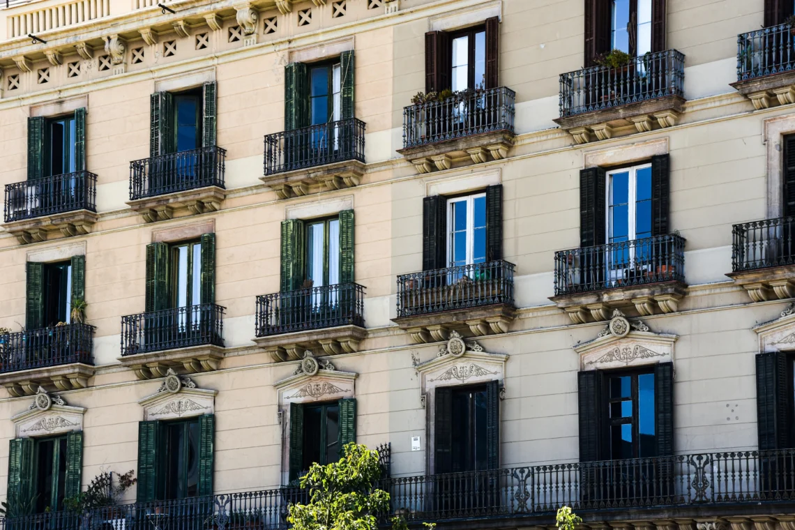 View of the façade of a classic residential building in Barcelona, featuring wrought-iron balconies, green and black shutters, and ornate architectural details