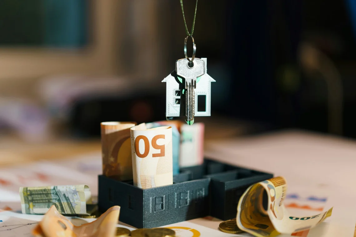 House key with home-shaped keychain hanging above euro banknotes in a miniature building model