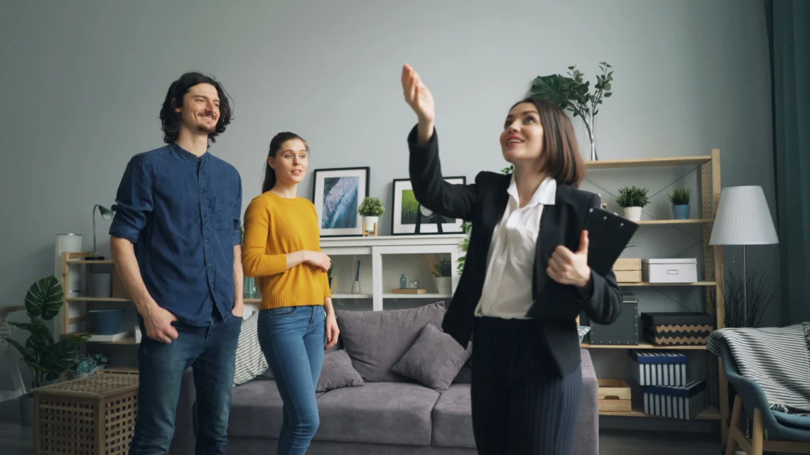 Un agent immobiliari somrient en un vestit fa gestos mentre mostra una sala d'estar moderna a una parella jove. L'habitació compta amb un sofà gris, obres d'art emmarcades, plantes i prestatgeries, creant un interior acollidor i elegant.