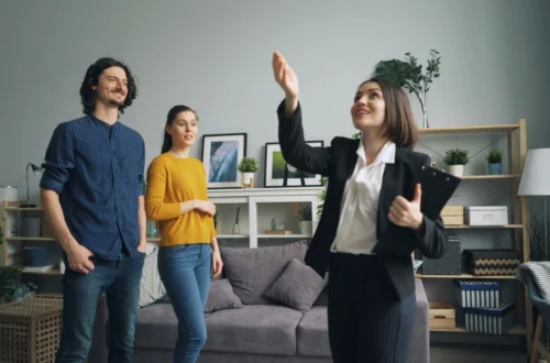 Un agent immobiliari somrient en un vestit fa gestos mentre mostra una sala d'estar moderna a una parella jove. L'habitació compta amb un sofà gris, obres d'art emmarcades, plantes i prestatgeries, creant un interior acollidor i elegant.