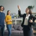 Un agent immobiliari somrient en un vestit fa gestos mentre mostra una sala d'estar moderna a una parella jove. L'habitació compta amb un sofà gris, obres d'art emmarcades, plantes i prestatgeries, creant un interior acollidor i elegant.
