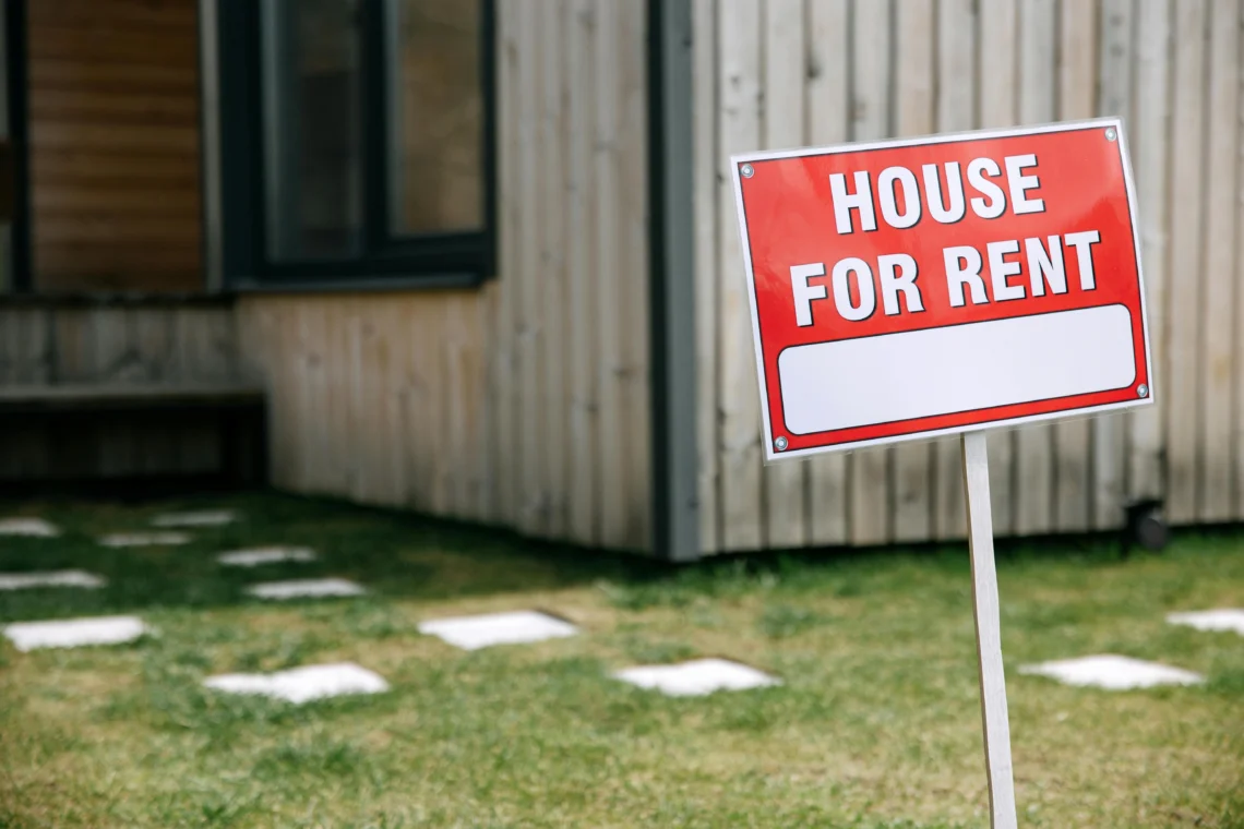 A red “House for Rent” sign placed in front of a wooden house with a well-kept garden and paved walkways.