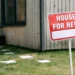 A red “House for Rent” sign placed in front of a wooden house with a well-kept garden and paved walkways.