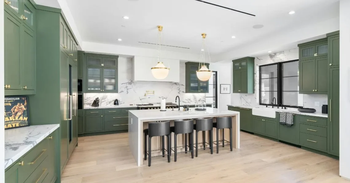 Amplia cocina moderna con muebles verdes, encimeras y salpicadero en mármol blanco, isla central con taburetes negros y lámparas colgantes doradas sobre suelos de madera clara.