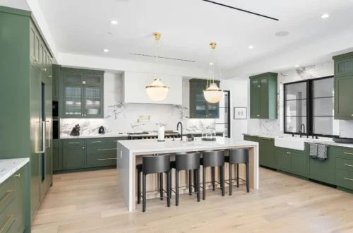 Amplia cocina moderna con muebles verdes, encimeras y salpicadero en mármol blanco, isla central con taburetes negros y lámparas colgantes doradas sobre suelos de madera clara.