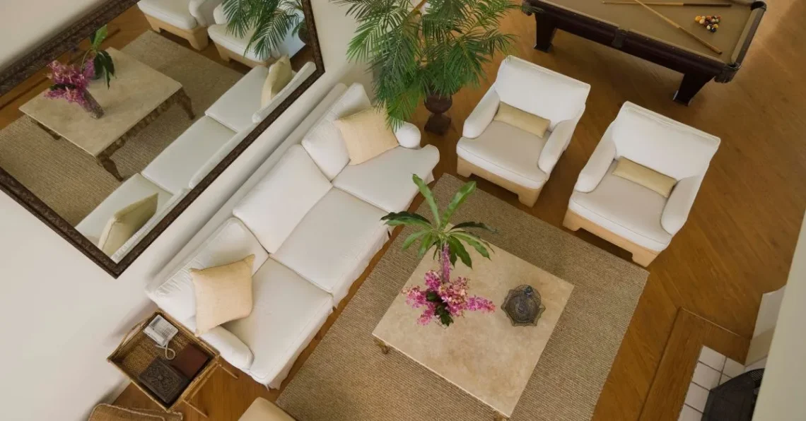 Vista aérea de una sala de estar elegante, con sofás blancos, mesas de centro de mármol, plantas decorativas y un espejo grande, todo en un ambiente cálido y acogedor.