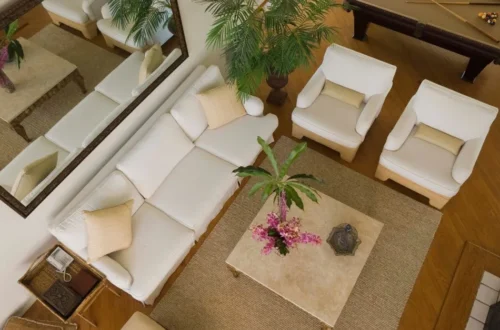 Vista aérea de una sala de estar elegante, con sofás blancos, mesas de centro de mármol, plantas decorativas y un espejo grande, todo en un ambiente cálido y acogedor.