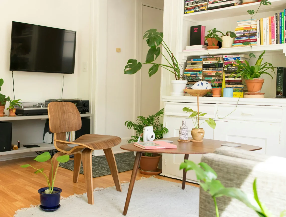 Sala de estar acogedora y luminosa con una estantería llena de libros, varias plantas, una mesa de centro de madera y una televisión en la pared.