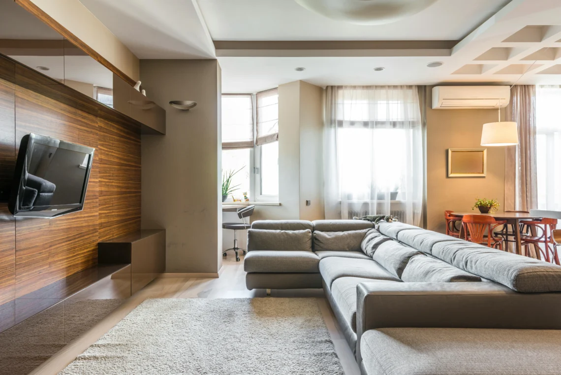 Vista de un salón moderno con sofá en forma de L, pared de madera con televisor, comedor al fondo y luz natural entrando por las ventanas.